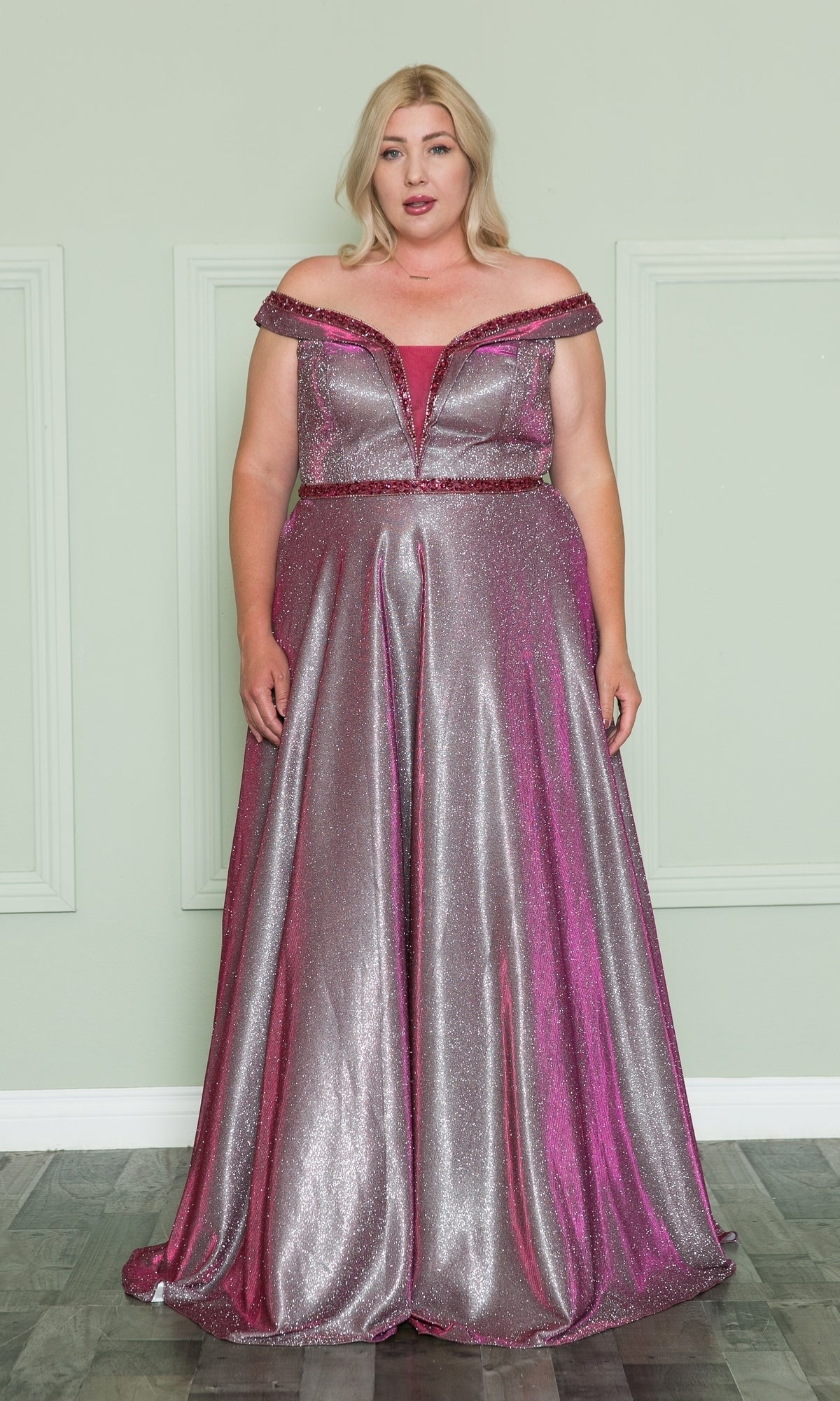 A woman stands in front of a light green wall wearing the Plus-Size Long Glitter-Knit Prom Dress W1096, an off-the-shoulder silver and pink gown with glitter accents and a floor-length skirt.